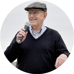 older gentleman in hat and glasses smiling into microphone